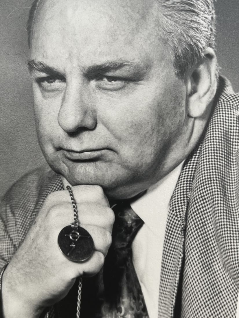 Studio portrait of Ted Owens, hand at his chin, holding a medallion on a chain.